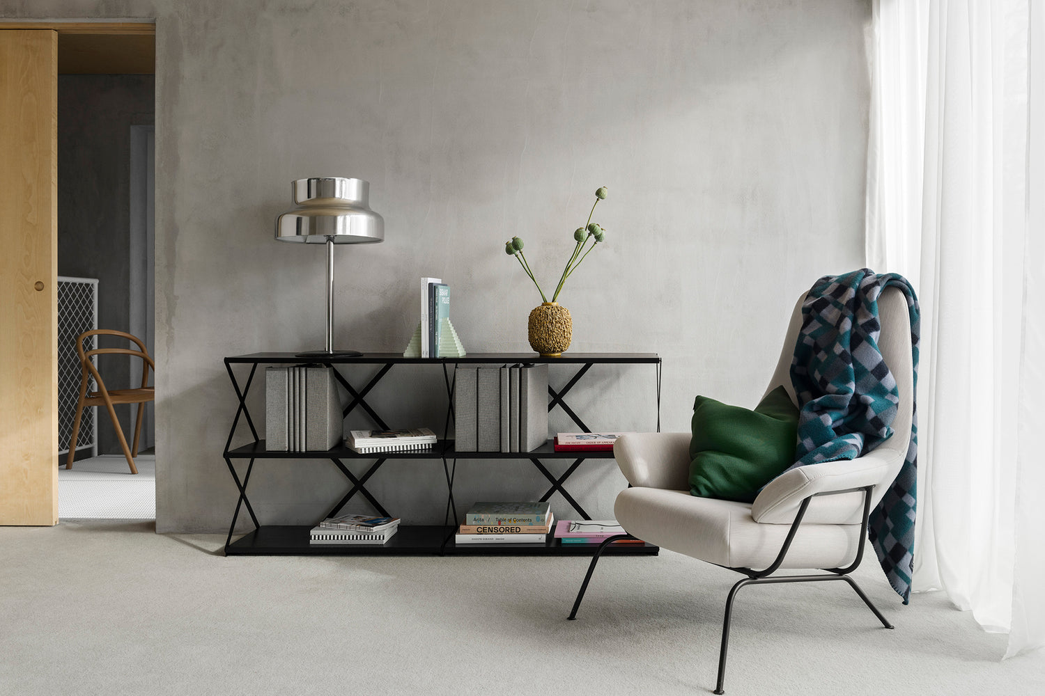 A living room / lounge scene featuring a Hai Lounge Chair in Shell with a Vienna Throw draped over it, and a Lift Shelf 6 in Black behind.