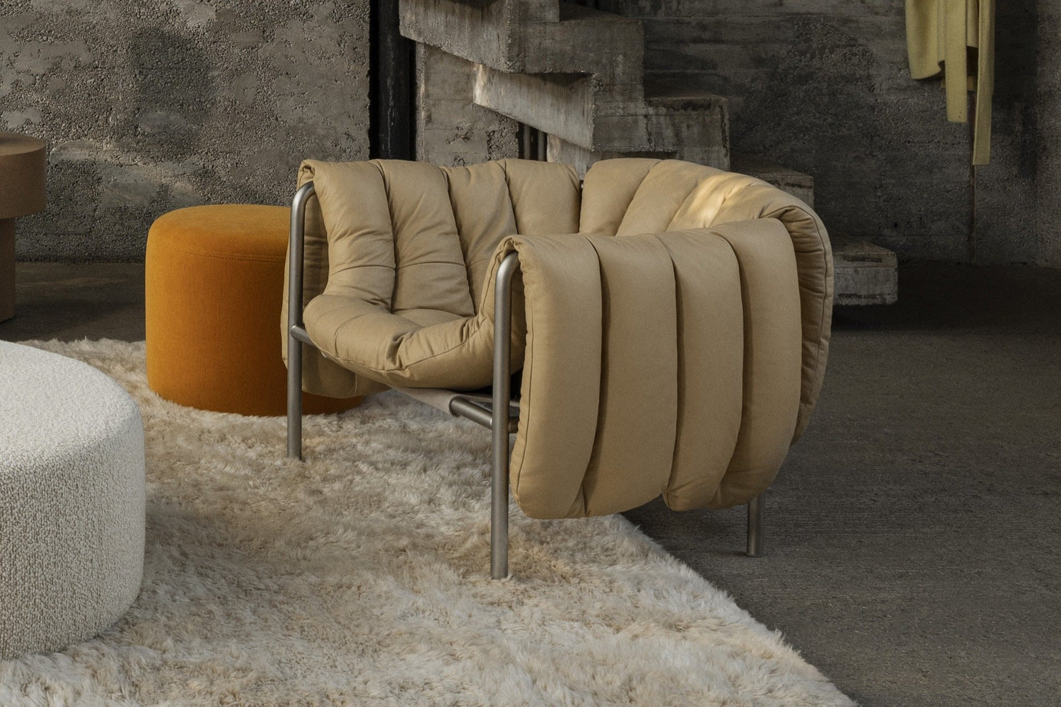 A living room scene featuring a Puffy Lounge Chair in Sand Soft Leather / Stainless Steel on top of a Monster Rug in Beige / Off-White. In the background is a Bon Pouf Round in Ochre and a Bon Pouf Round Large in Eggshell.