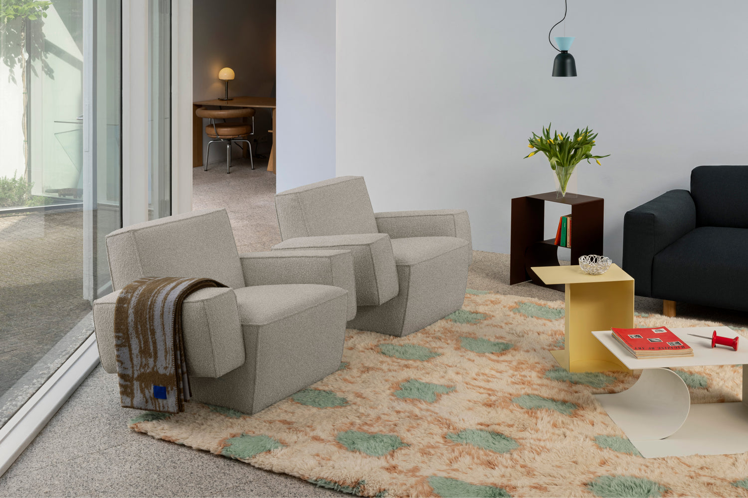 A lifestyle image of a living room / lounge scene featuring Hunk Lounge Chair with Armrests, Glitch Throw, Monster Rug, Alphabeta Pendant Light, and Glyph Side Tables.