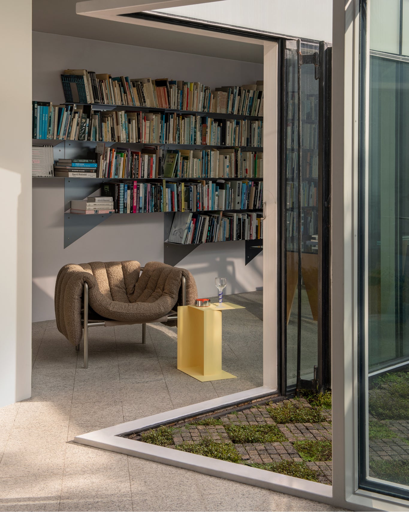 A lifestyle image of a library featuring Puffy Lounge Chair in Sawdust / Stainless beside a Glyph Side Table Gamma in Wax Yellow.