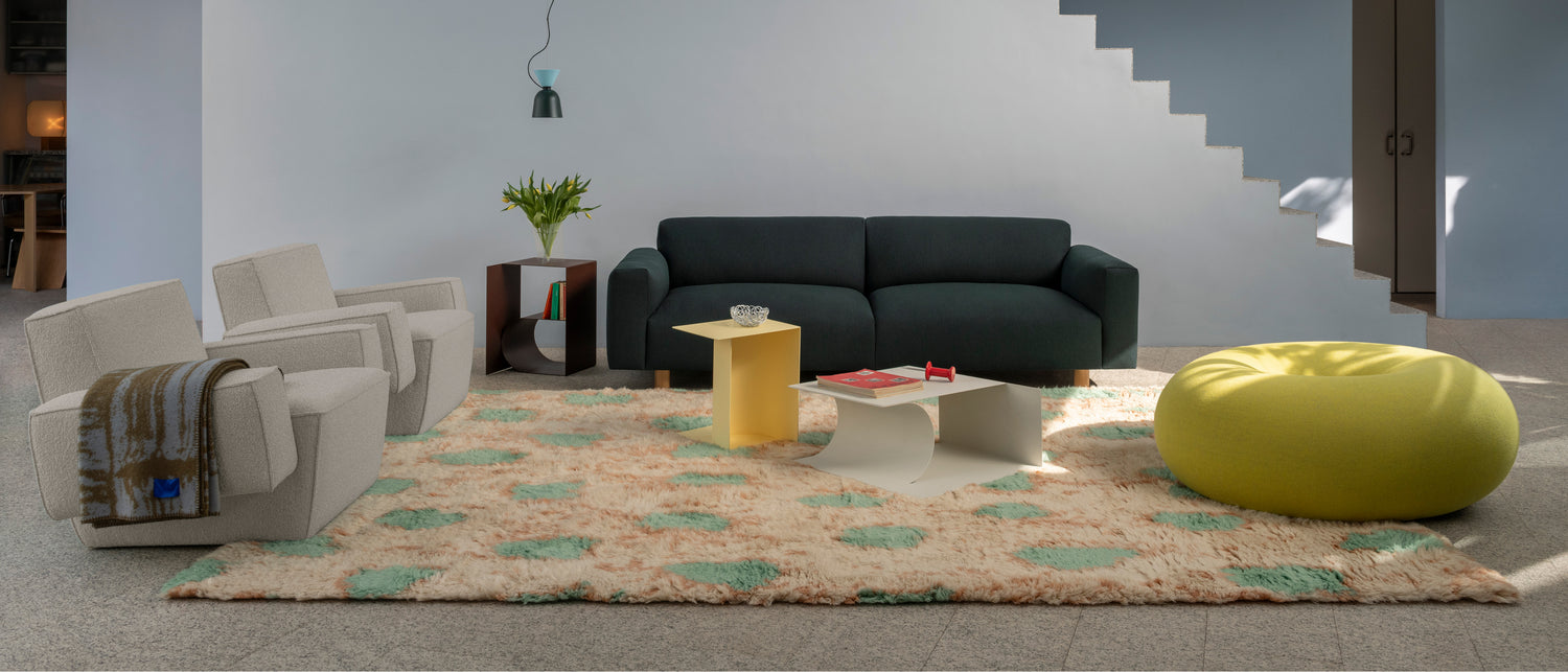 A lifestyle image of a living room scene featuring Hunk Lounge Chair with Armrests, Glitch Throw, Monster Throw, Glyph Side Table, Boa Pouf, and Alphabeta Pendant Light.