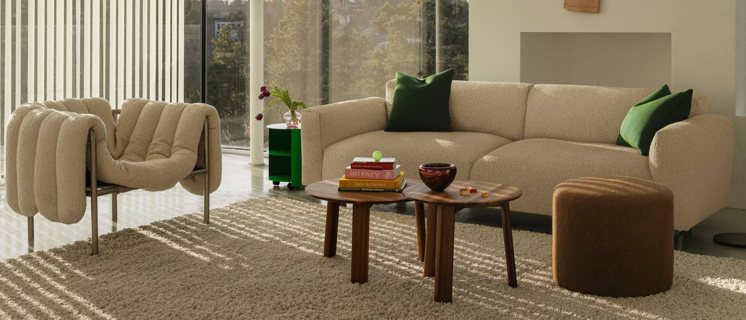 A living room scene featuring a Puffy Lounge Chair in Eggshell / Stainless Steel, Hide Side Table Pure Green, Koti 3-seater Sofa, Alle Coffee Tables Set of 2 Walnut, Bon Pouf Round Brown, and an Alphabeta Floor Lamp.
