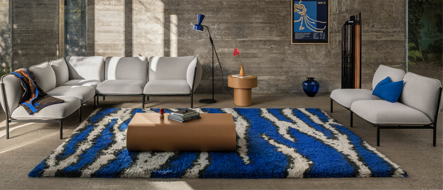 A living room scene featuring Kumo Sofas in Porcelain, Monster Rug Ultramarine Blue / Off-White, Arch Throw Black / Brown / Blue, Stump Coffee Table Medium Natural, Alphabeta Floor Lamp Blue, Stump Side Table Natural, and Storm Cushion Large Sky.